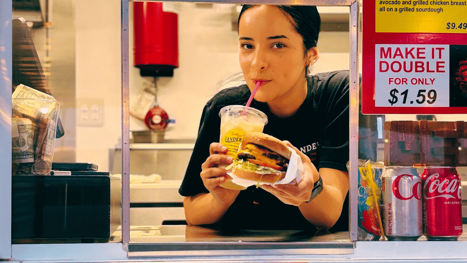 Woman sipping a drink with a burger ad behind her at a food stand.